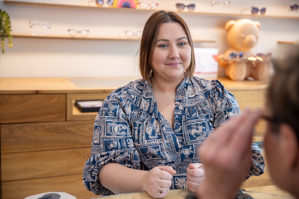 A woman sits at a desk, smiling, possibly assisting a customer in an optical shop. Glasses are displayed in the background - Carrboro Eye Doctor A woman sits at a desk, smiling, possibly assisting a customer in an optical shop. Glasses are displayed in the background - Carrboro Eye Doctor