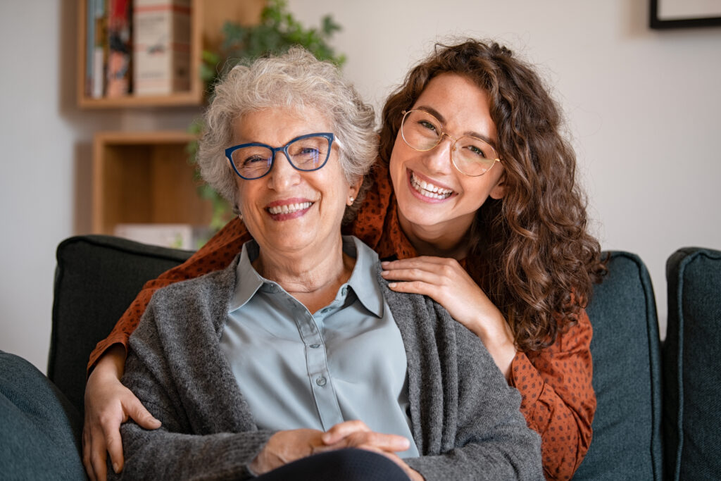 A happy elderly woman and a young woman, both wearing glasses, smiling while sitting together on a couch - Carrboro Eye Doctor A happy elderly woman and a young woman, both wearing glasses, smiling while sitting together on a couch - Carrboro Eye Doctor